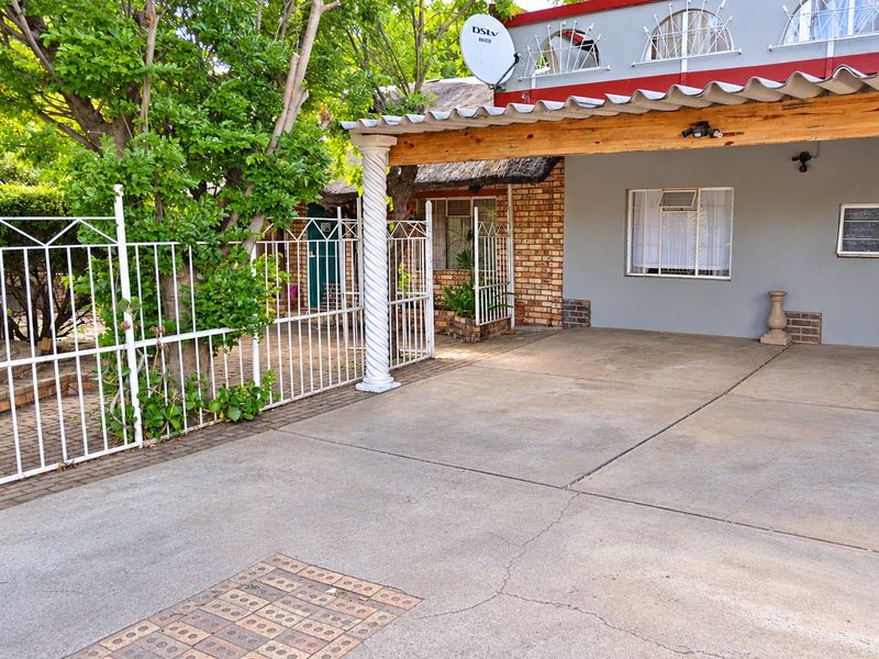 Front exterior view featuring a concrete driveway, covered carport area, decorative column, and secure metal fencing with brick and painted wall finishes.