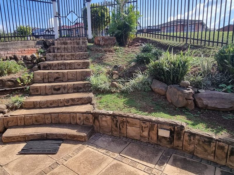 A landscaped garden area featuring stone retaining walls, paved patio space, and stone steps leading to a metal gated entrance.