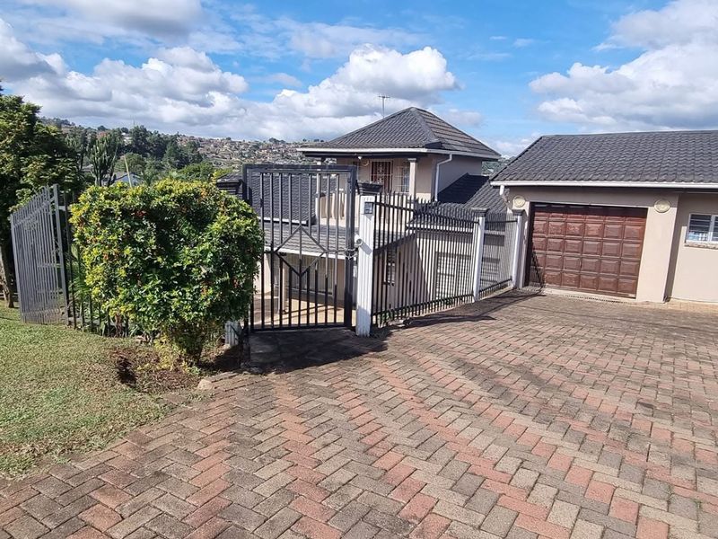 Front exterior view of the property featuring a paved driveway, secure metal gate, perimeter fencing, and a double-story house with a tiled roof.