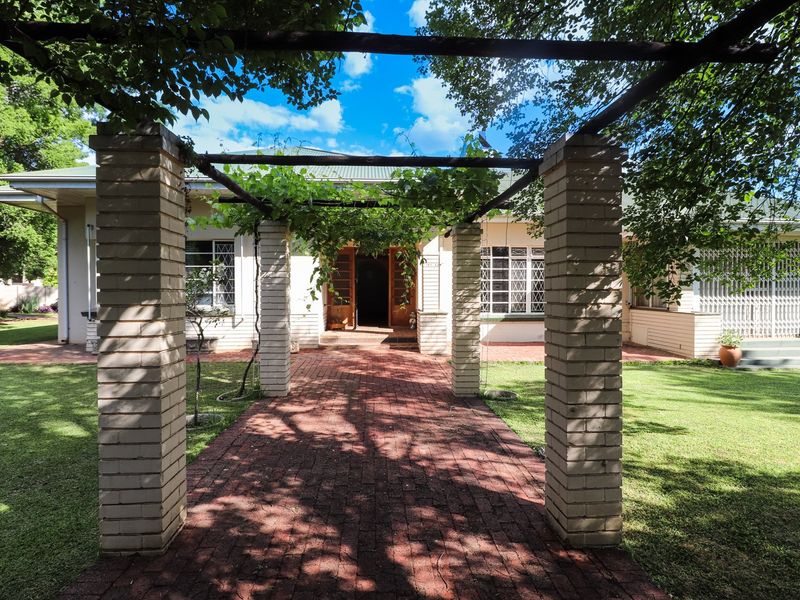 A charming front entrance featuring a brick-paved walkway framed by a vine-covered timber pergola, leading to the main house facade with security gates.