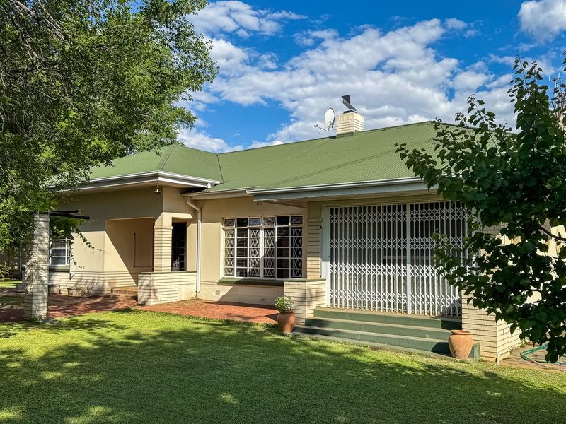 Rear view of a single-story house featuring a green corrugated metal roof, brick exterior, and a paved patio area overlooking a garden.