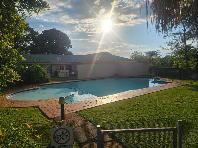 A communal swimming pool area featuring brick paving, surrounding lawn, and boundary walling, set within a landscaped garden environment.