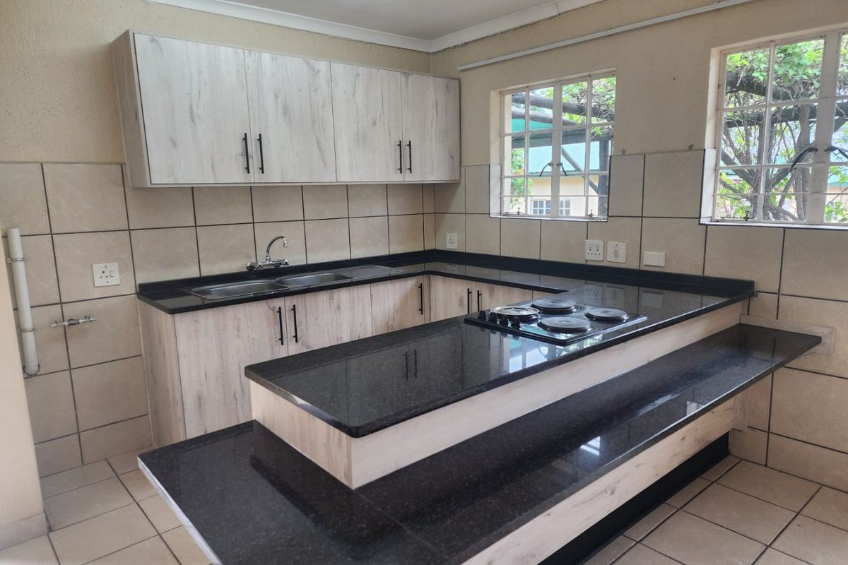 A clean, functional kitchen featuring light wood-finish cabinetry, dark granite countertops, a built-in electric hob, and tiled splashbacks with ample natural light.
