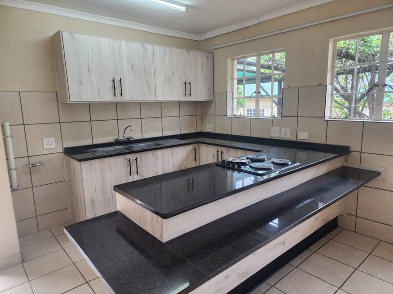A clean, functional kitchen featuring light wood-finish cabinetry, dark granite countertops, a built-in electric hob, and tiled splashbacks with ample natural light.
