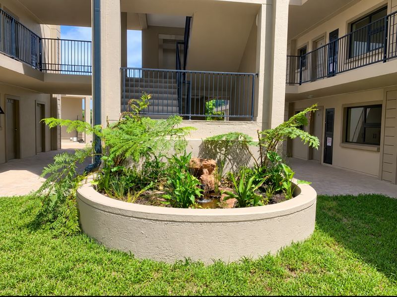 A well-maintained apartment complex courtyard featuring a central concrete planter with a water feature, surrounded by paved walkways and metal balustrades.