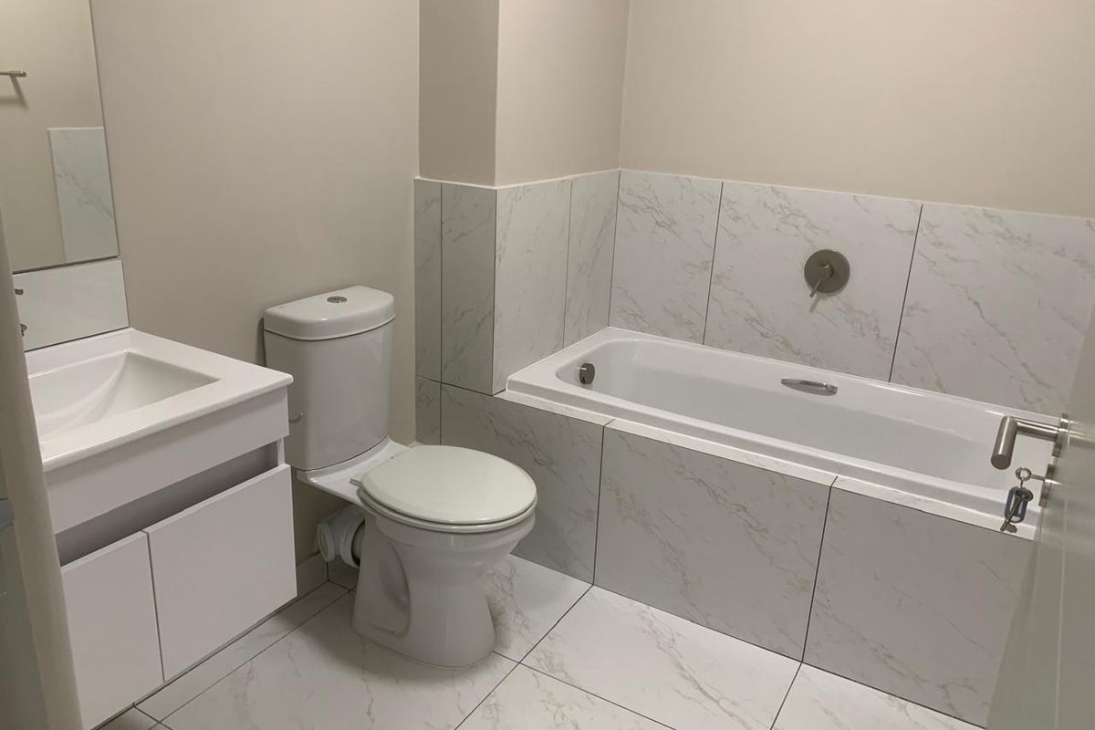 A modern, clean bathroom featuring a built-in vanity, toilet, and a bathtub with marble-look tiled surround and wall-mounted fixtures.