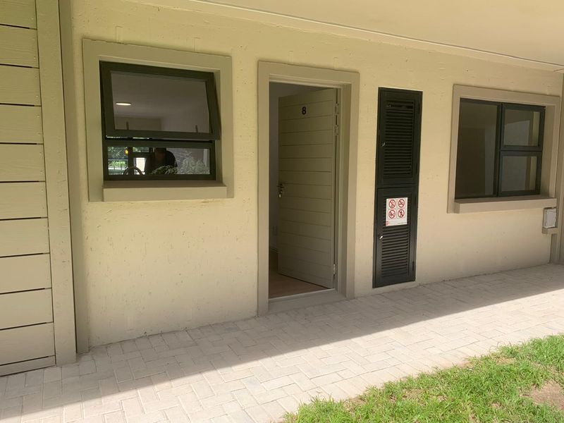 Exterior view of an apartment entrance featuring a paved walkway, aluminium window frames, and a secure louvered utility cupboard.