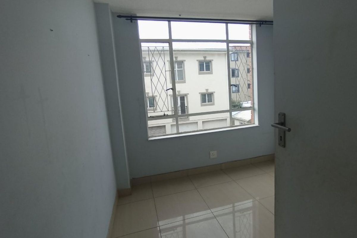 A bright, empty bedroom featuring neutral tiled flooring and a large window with security bars, offering a clean and versatile living space.