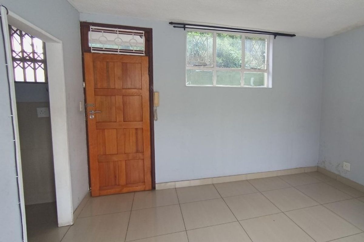 An empty living area featuring tiled flooring, a wooden entrance door with security gate, and a window with security bars.
