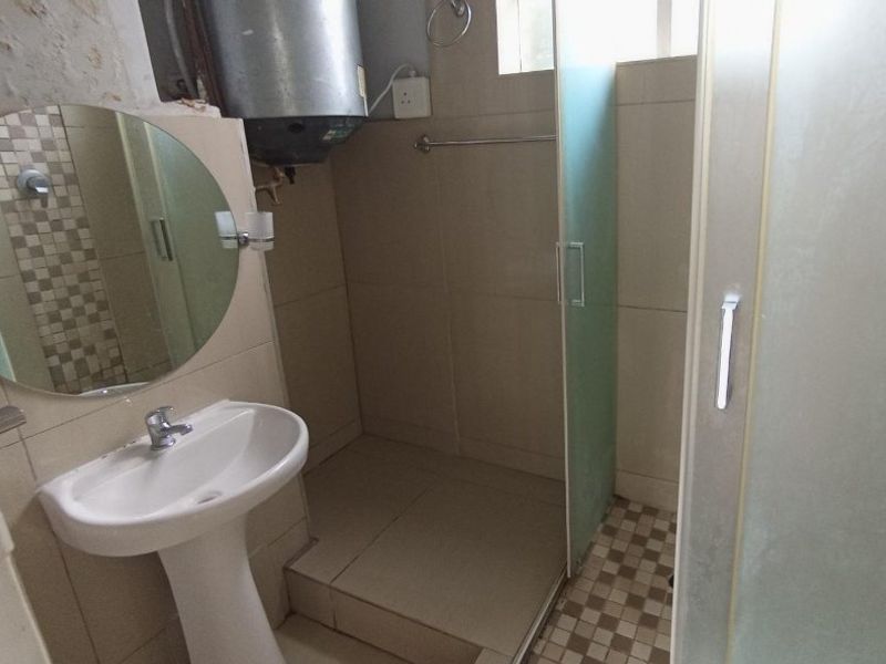 A compact bathroom featuring a pedestal basin, shower enclosure, and tiled finishes. The space includes an exposed electric geyser and requires minor cosmetic finishing.