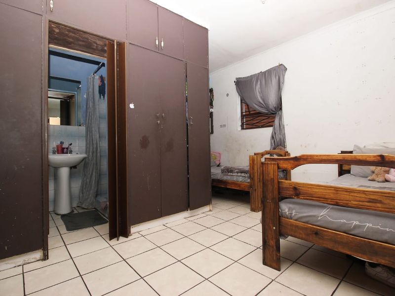 Bedroom featuring built-in cupboards, tiled flooring, and an en-suite bathroom with a pedestal basin. The room requires cosmetic updates to wall finishes and cabinetry.