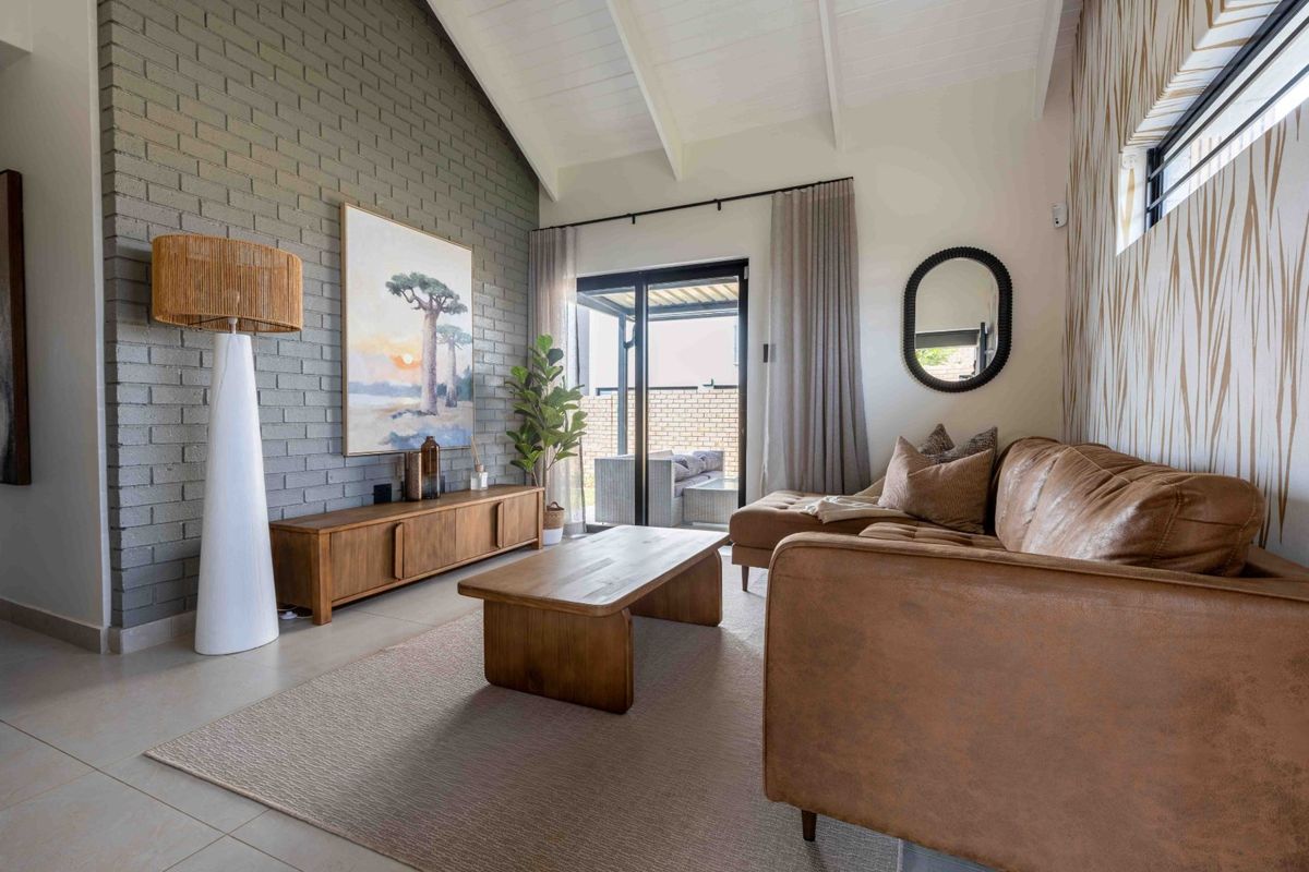 A bright living room featuring a vaulted ceiling with exposed rafters, a textured brick feature wall, and large sliding glass doors leading to the patio.