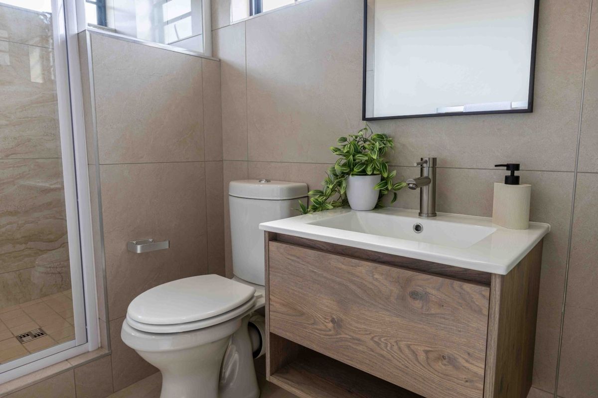 Modern bathroom featuring a wood-finish vanity cabinet, ceramic basin, toilet, and a glass-enclosed shower with neutral wall and floor tiling.