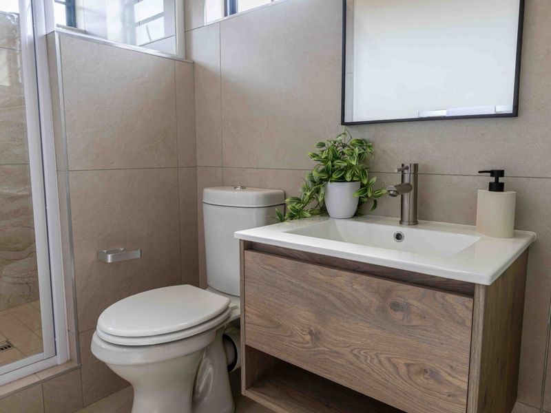Modern bathroom featuring a wood-finish vanity cabinet, ceramic basin, toilet, and a glass-enclosed shower with neutral wall and floor tiling.