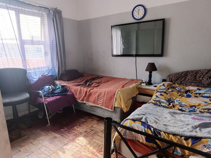 A bedroom featuring traditional parquet flooring and a window, currently utilized as a shared sleeping space with visible wear on wall finishes.