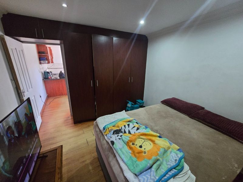 A bedroom featuring built-in wooden cupboards, laminate flooring, and recessed downlighting, with an adjacent view into the kitchen area.