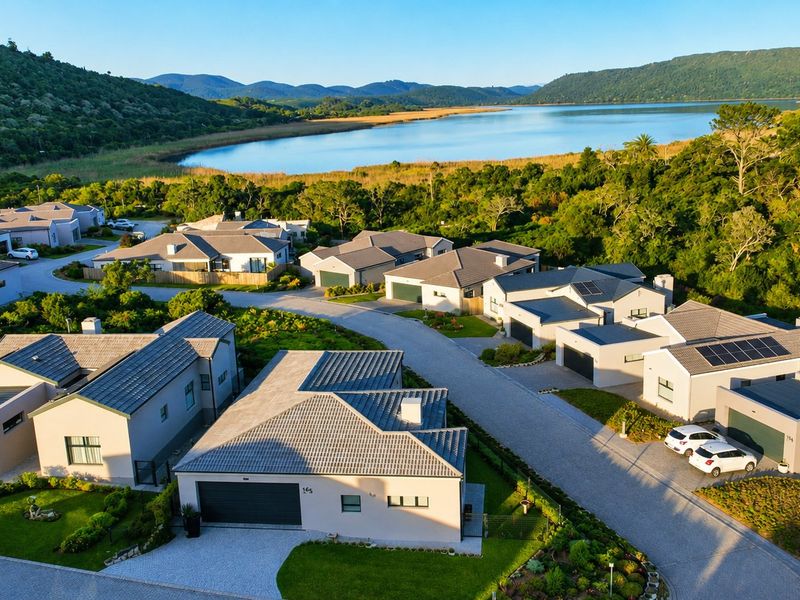 Aerial view of a modern residential estate featuring contemporary homes with tiled roofs, solar panels, and landscaped gardens, overlooking a scenic lake and mountains.