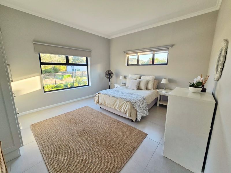 A bright and spacious bedroom featuring neutral tiled flooring, modern aluminium window frames with roller blinds, and built-in storage cabinetry.