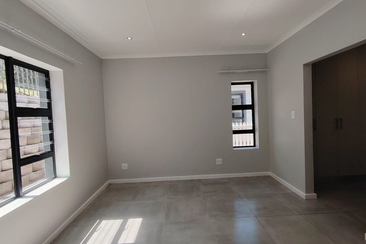 A bright, empty bedroom featuring neutral grey walls, large aluminium-framed windows, modern tiled flooring, and built-in cupboard storage.