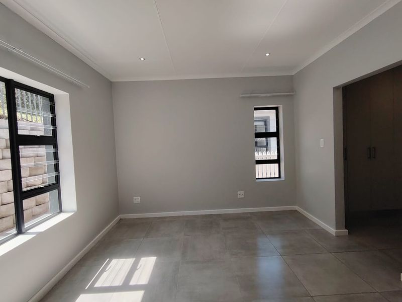 A bright, empty bedroom featuring neutral grey walls, large aluminium-framed windows, modern tiled flooring, and built-in cupboard storage.
