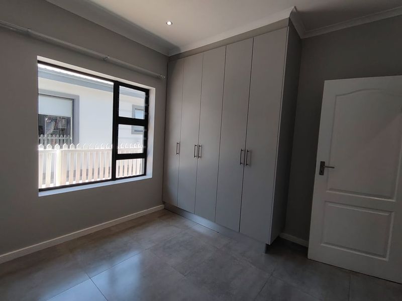 A clean, modern bedroom featuring neutral grey walls, large built-in cupboards, tiled flooring, and an aluminium window frame.