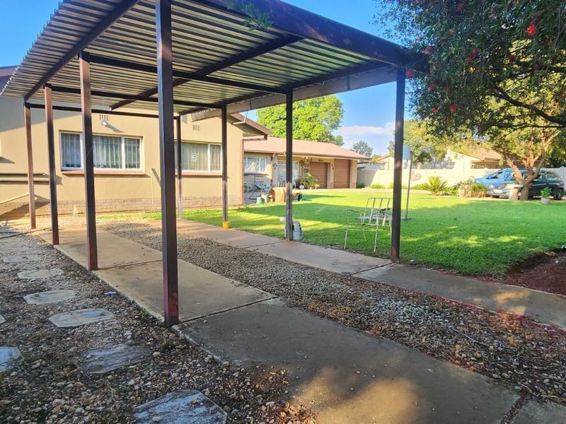 Rear exterior view featuring a metal carport structure, concrete driveway, and a spacious lawn area with established garden landscaping.