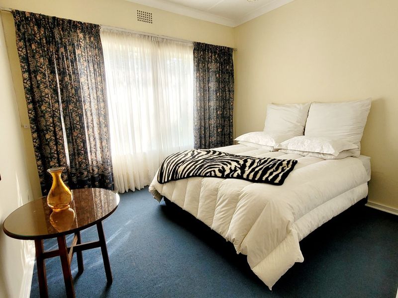 A standard bedroom featuring neutral walls, blue carpeting, and large windows with layered curtains, providing a functional and well-lit living space.