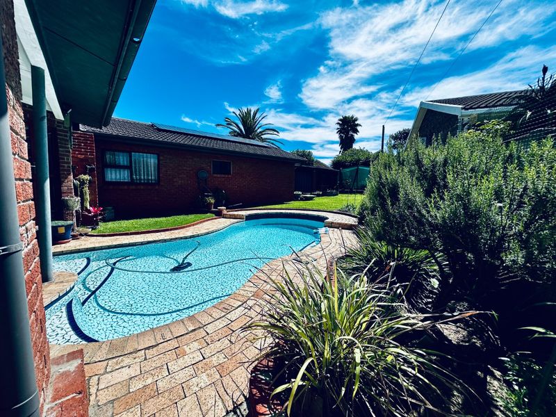 A well-maintained backyard featuring a curved swimming pool with brick paving surround, set against a brick house exterior with solar panels.