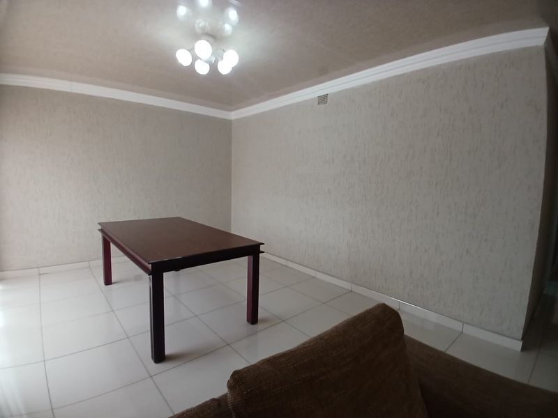 A spacious living area featuring neutral textured wallpaper, white tiled flooring, and decorative ceiling cornicing.
