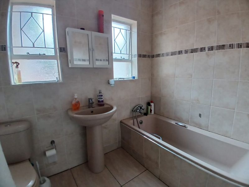 A functional bathroom featuring a pedestal basin, bathtub, and toilet with neutral wall tiling and security-barred windows.