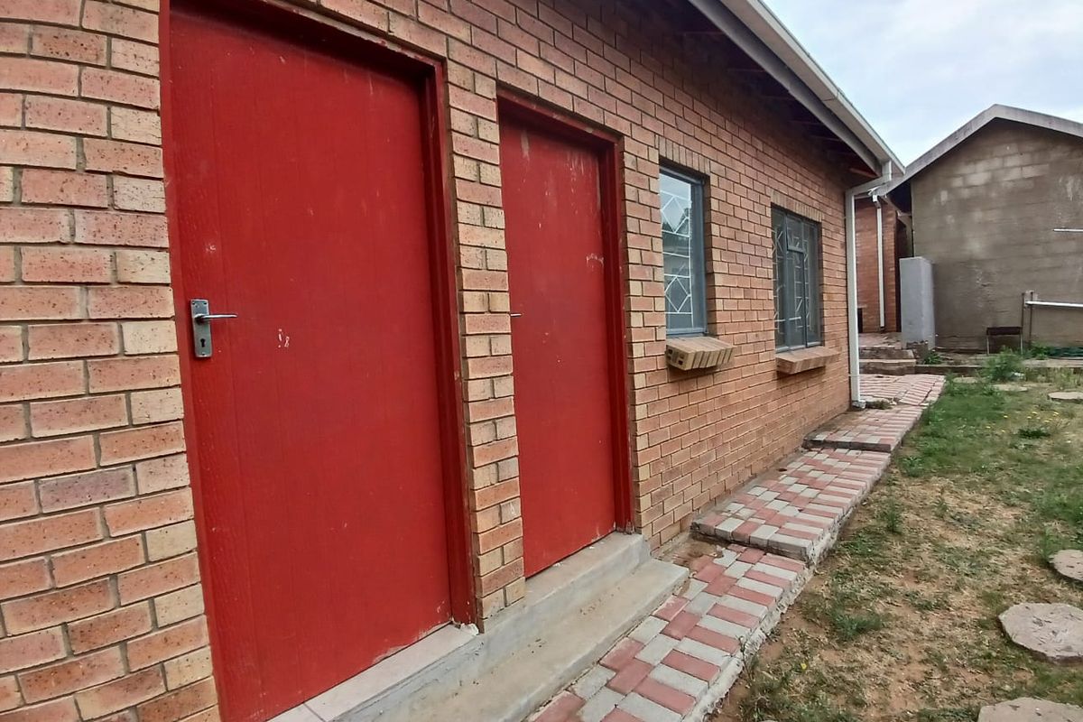 Rear exterior view of a face brick house featuring red doors, security-barred windows, and a paved walkway along the building perimeter.