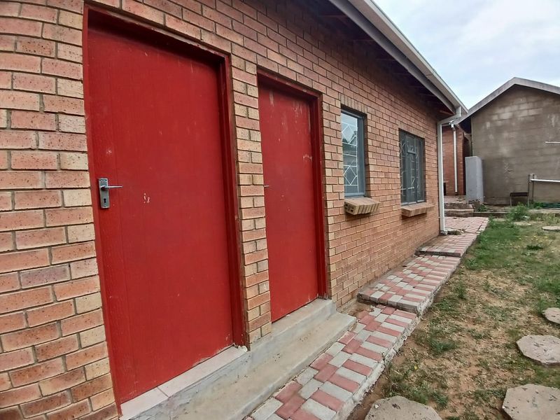 Rear exterior view of a face brick house featuring red doors, security-barred windows, and a paved walkway along the building perimeter.