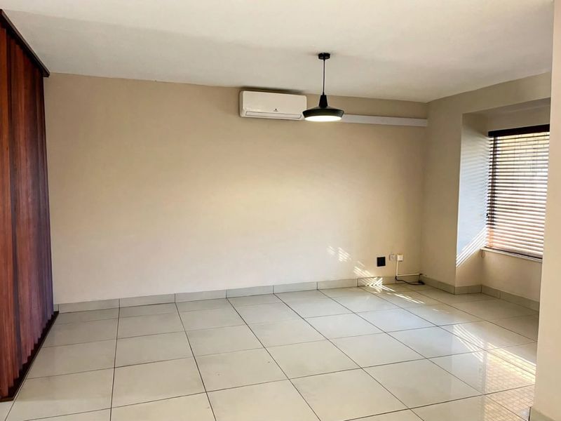 A bright, empty living area featuring neutral tiled floors, a wall-mounted air conditioning unit, and natural light filtering through venetian blinds.