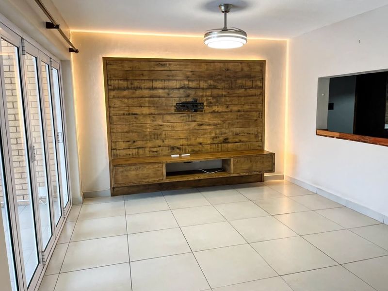 A bright, modern living area featuring a custom built-in wooden media console, ambient LED lighting, tiled floors, and large sliding doors leading outdoors.