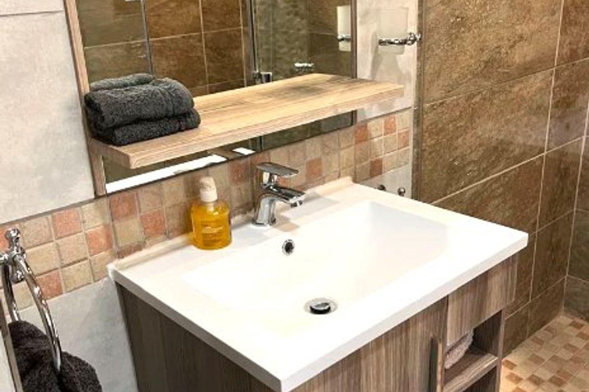 A modern bathroom featuring a wood-finish vanity cabinet with a white basin, wall-mounted mirror, and a tiled shower enclosure.