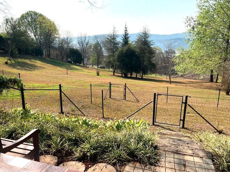A spacious garden area featuring wire mesh fencing, a metal gate, and a paved walkway, overlooking a large grassy landscape with mature trees.
