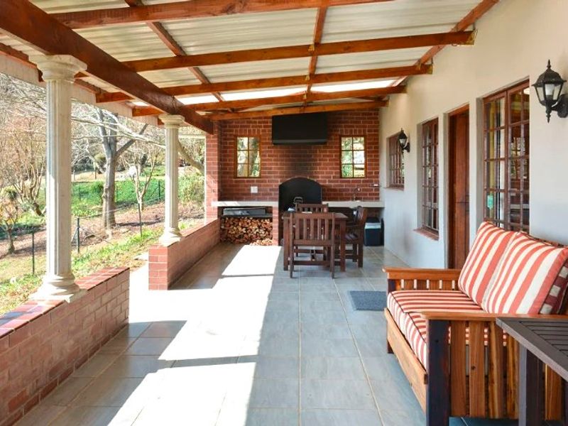A spacious covered patio featuring a built-in brick braai, tiled flooring, and timber roof structure, overlooking the garden area.