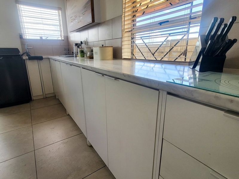 A functional scullery area featuring white cabinetry, marble-look countertops, tiled flooring, and a tiled backsplash with natural light from windows.