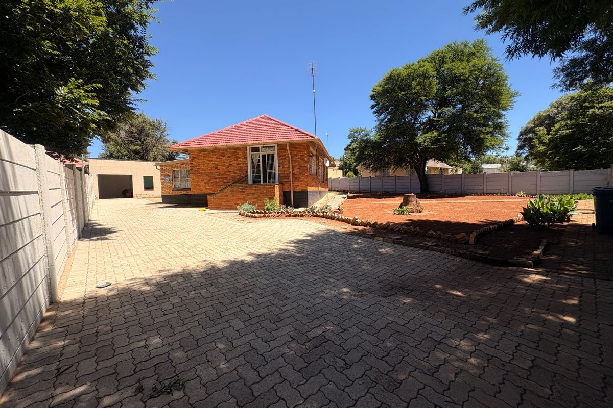 Rear view of a brick house featuring a large paved driveway, detached garage, and a spacious garden area enclosed by precast concrete walls.
