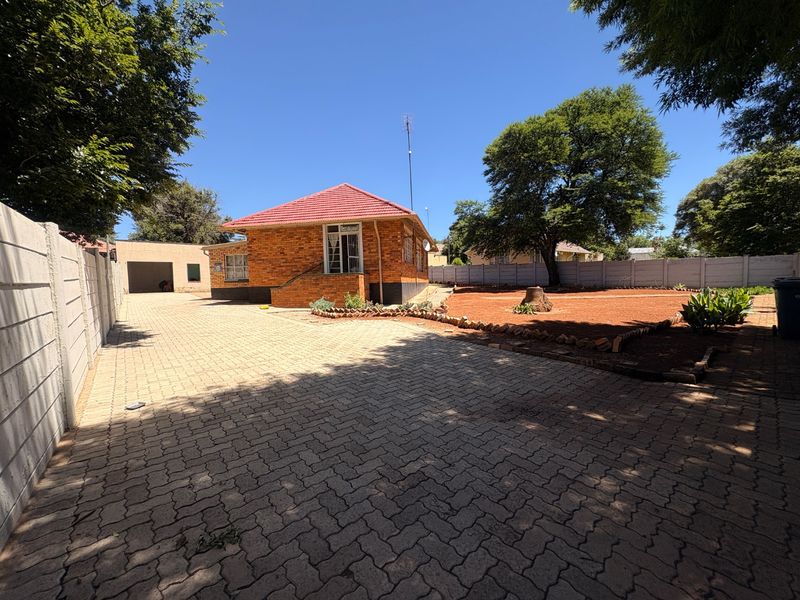 Rear view of a brick house featuring a large paved driveway, detached garage, and a spacious garden area enclosed by precast concrete walls.