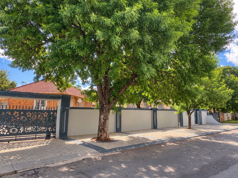 Front view of the property featuring a secure perimeter wall, decorative metal gate, and paved driveway, complemented by mature trees and a tiled roof.