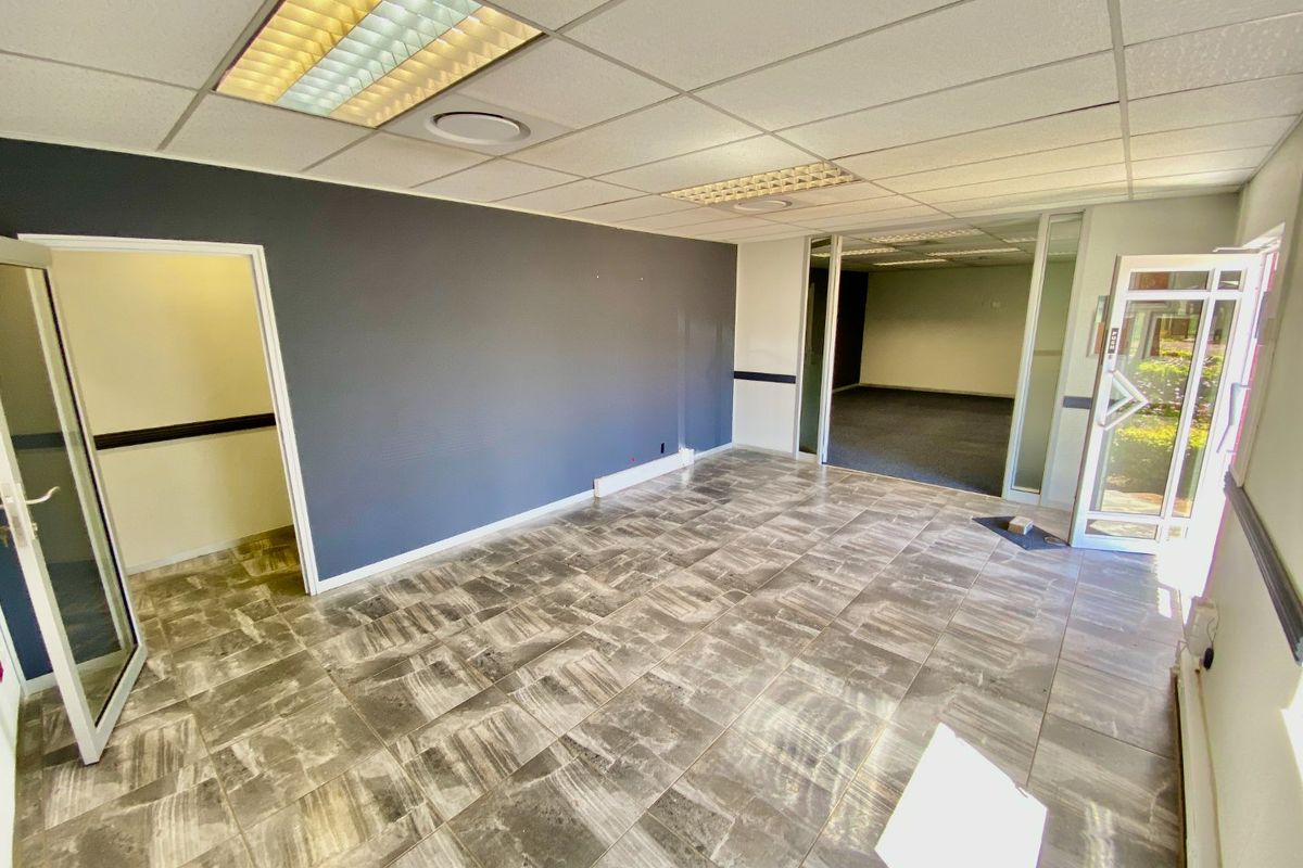 An empty commercial office space featuring tiled flooring, a suspended ceiling with recessed lighting, and a glass partition wall leading to an adjacent area.