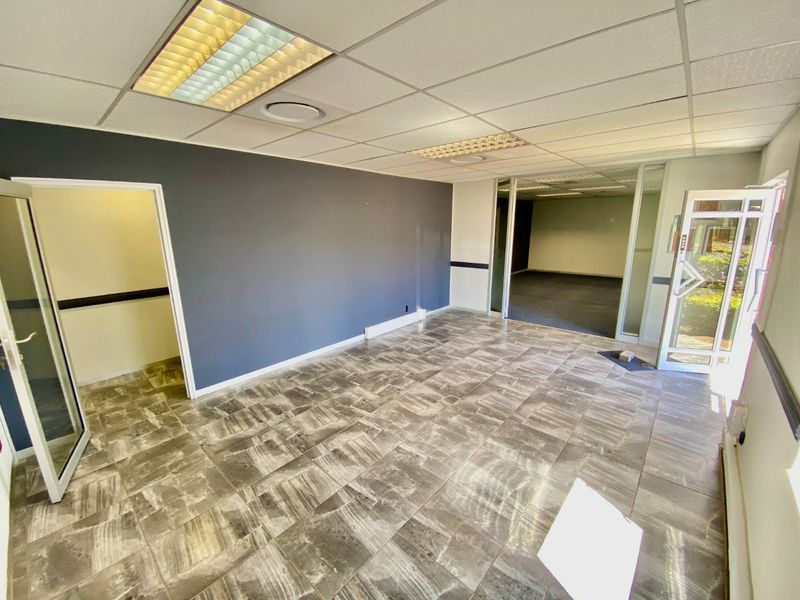 An empty commercial office space featuring tiled flooring, a suspended ceiling with recessed lighting, and a glass partition wall leading to an adjacent area.
