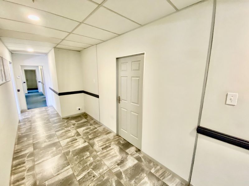A clean, well-lit commercial hallway featuring tiled flooring, a suspended ceiling, and a neutral color palette with a dado rail.