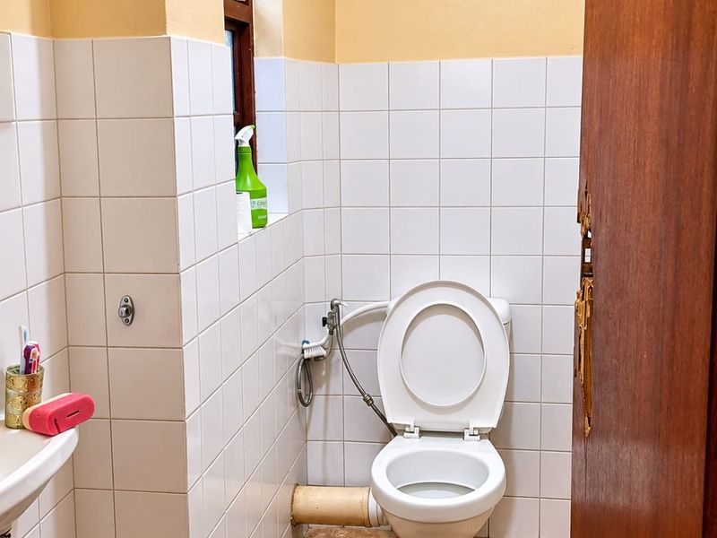 A functional bathroom featuring a standard toilet with bidet spray, wall tiling, and a washbasin, showing signs of wear and dated finishes.