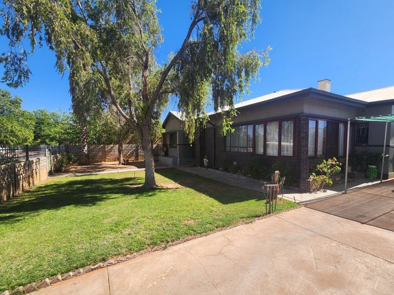 Rear view of the property featuring a brick facade, metal roof, paved driveway, and a well-maintained garden area with mature trees.