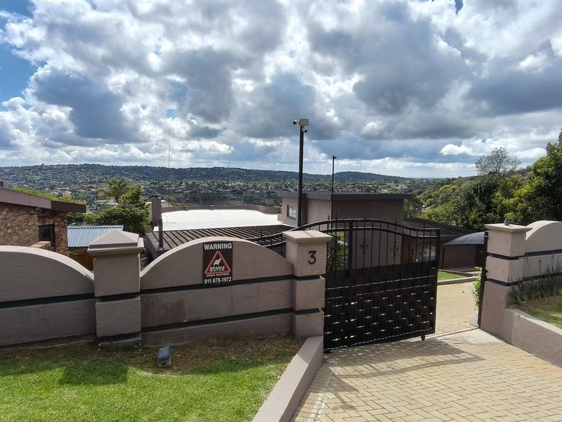 Front entrance view featuring a paved driveway, secure wrought iron gate, and masonry boundary wall with an elevated outlook over the surrounding area.