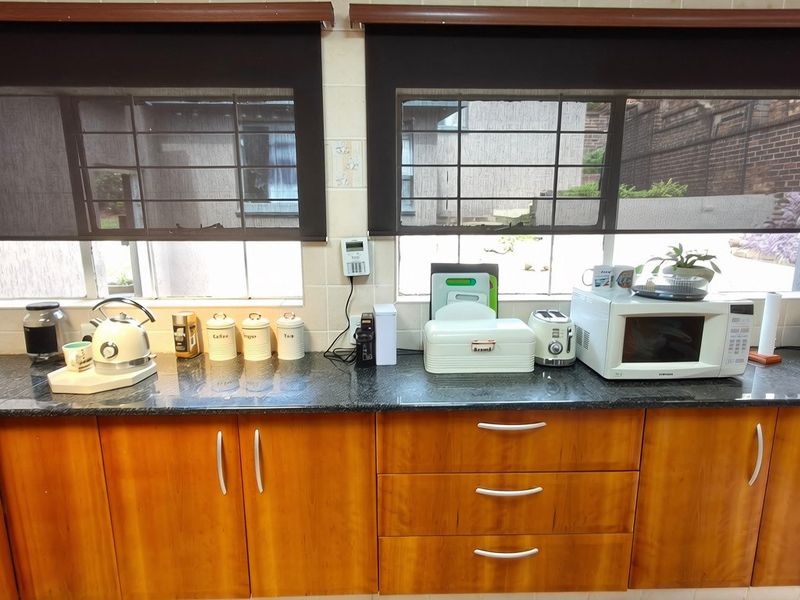 A functional kitchen area featuring granite countertops, warm-toned wooden cabinetry, and roller blinds, providing a practical workspace with integrated alarm system controls.