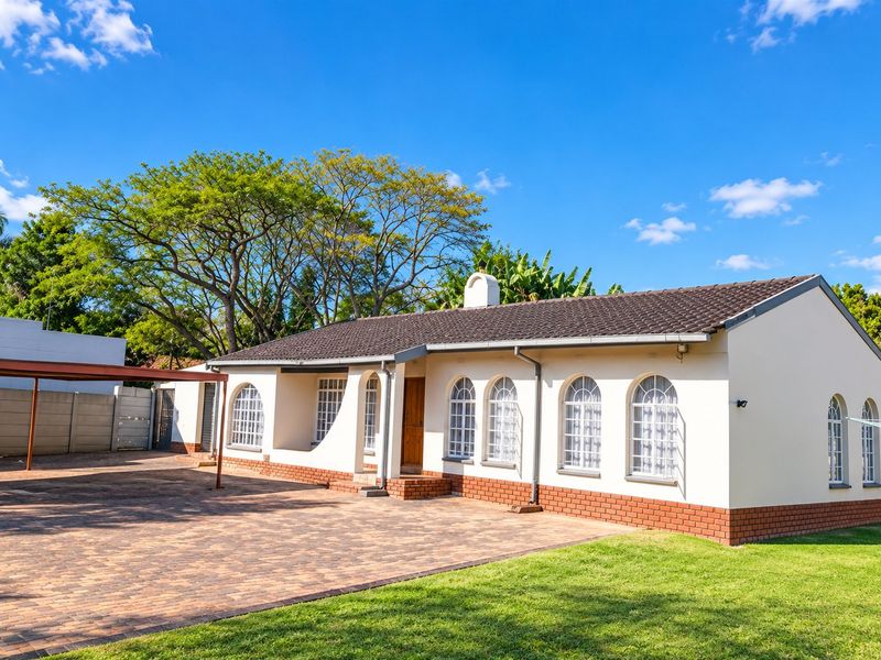 A well-maintained single-story house featuring a tiled roof, arched windows, brick detailing, and a spacious paved driveway with an attached carport.