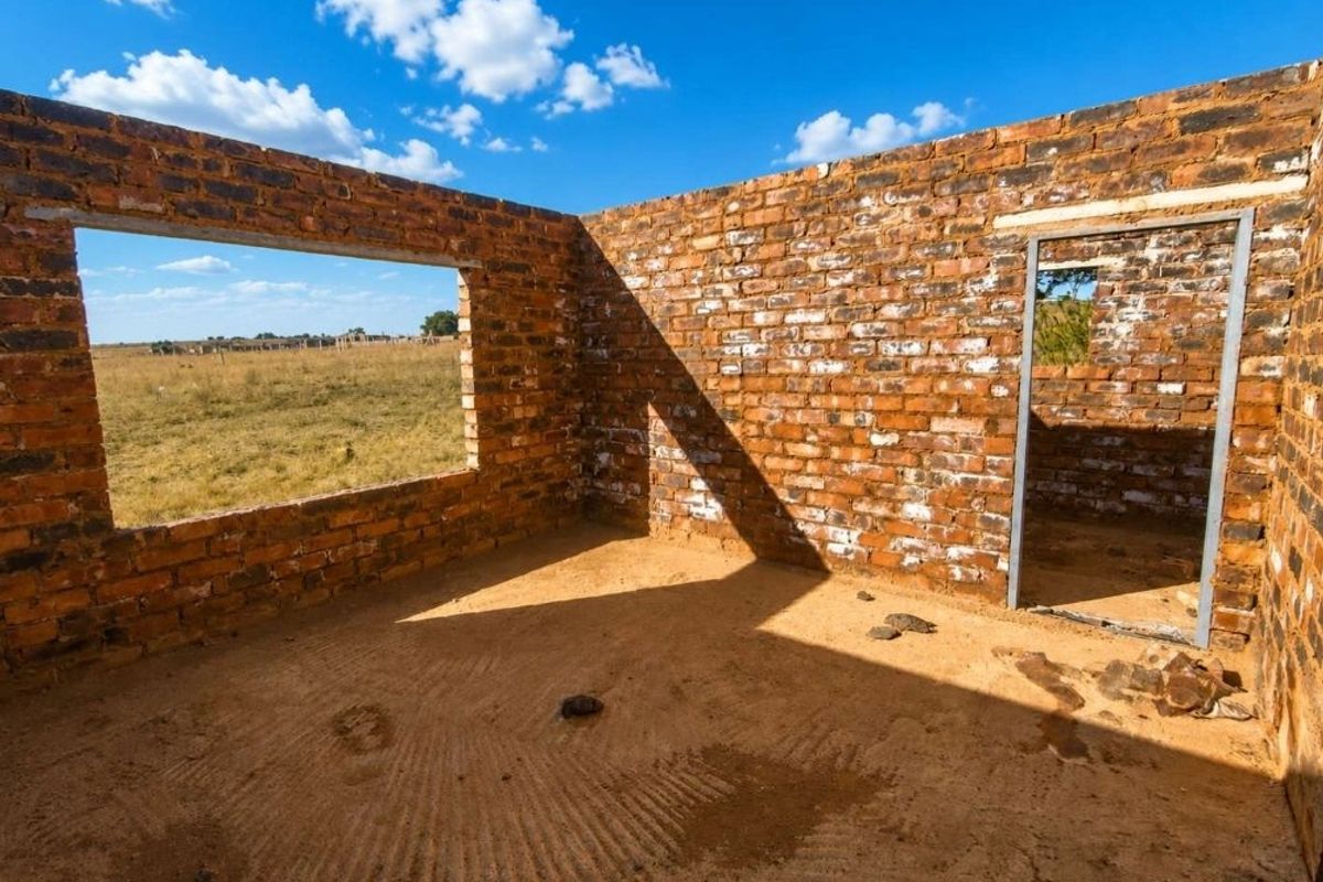 An unfinished construction site showing exposed brick walls, a metal door frame, and an open window frame on a rural farm property.